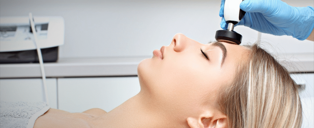 Woman receiving a non-invasive facial skin-tightening treatment using a handheld aesthetic device on the forehead, performed by a clinician wearing blue medical gloves in a professional clinic setting.