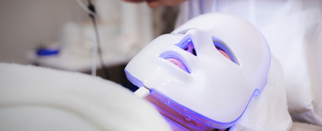 A client lying down wears a white LED light therapy mask glowing with blue-purple light during a professional skincare treatment.