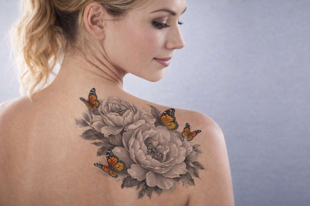 A close-up view of a woman's back showcasing an intricate tattoo featuring large gray roses and vibrant orange and black butterflies, highlighting a blend of floral and nature-inspired art.