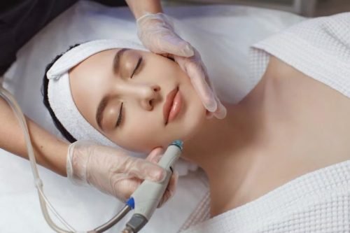 A young woman with her eyes closed receives a multi-step facial treatment on her chin using a handheld device that delivers or removes liquid, administered by a technician in gloves.